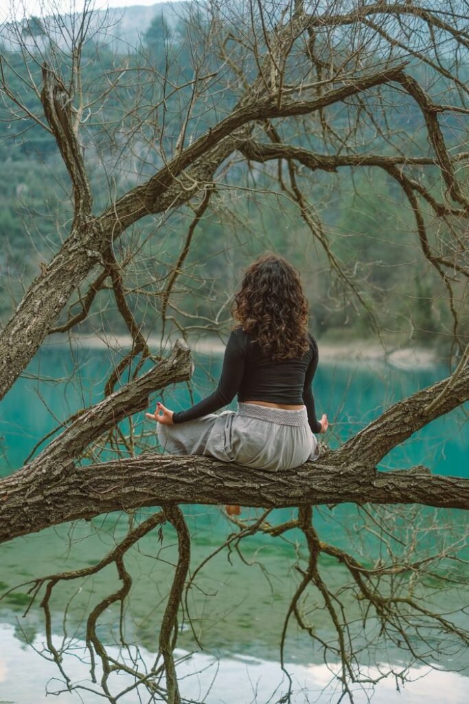 A woman practices meditation on a tree branch over a tranquil lake, embracing nature's serenity.